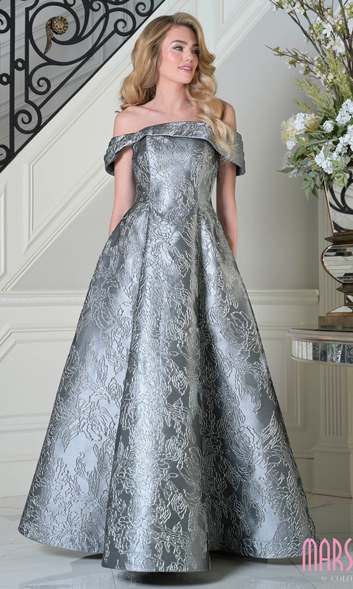 A woman wearing a charcoal off-the-shoulder Marsoni MV1395 long formal dress with a textured floral pattern stands indoors near a staircase and floral arrangement.